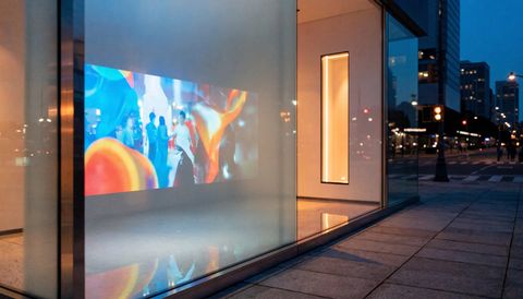 A commercial glass facade at dusk with a vivid brand video rear-projected onto a frosted smart-glass pane